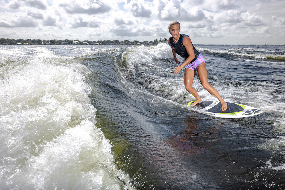 A woman in a life vest and purple shorts is wakesurfing on a lake behind a boat, riding the 2026 Phase 5 Aku v2 Wakesurf Board under a cloudy sky.