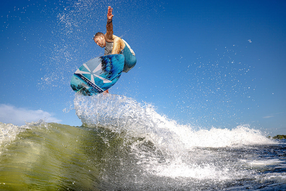 A surfer catches air above a wave, balancing on a blue and white Phase 5 Diamond Turbo Wakesurf Board 2026 with one arm raised, sunlight glinting off the water under a clear blue sky.
