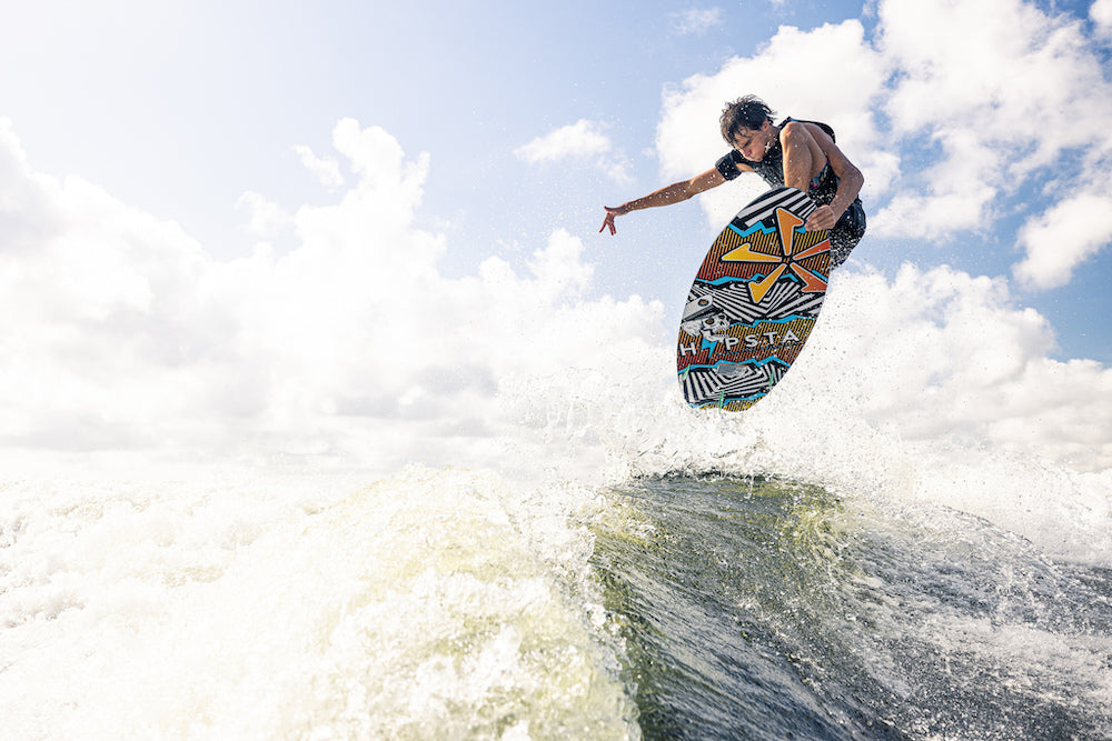 Someone rides the Phase 5 Hypsta v2 Wakesurf Board 2026 above a wave, sunlight gleaming under a partly cloudy sky.