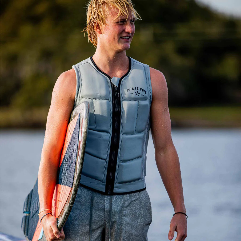 A young adult male is wearing a grey phase 5 comp vest while holding a wakesurf board by phase 5 under his arm