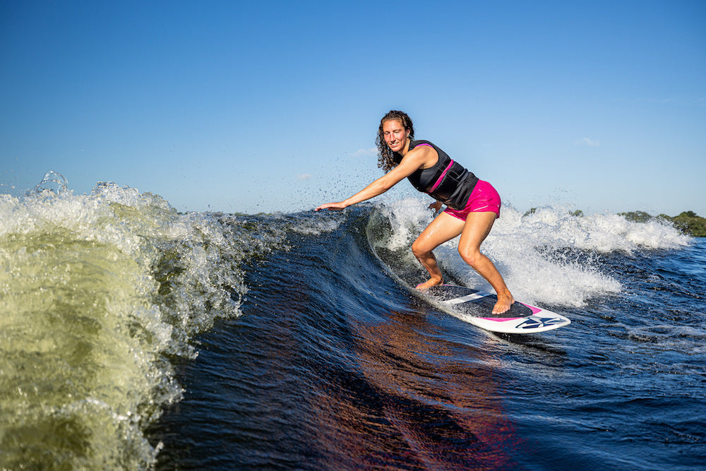 A woman in a black vest and pink shorts rides the Phase 5 Rush Wakesurf Board 2026 by Phase 5 under a clear blue sky, looking focused and balanced as she highlights the board’s lightweight performance.