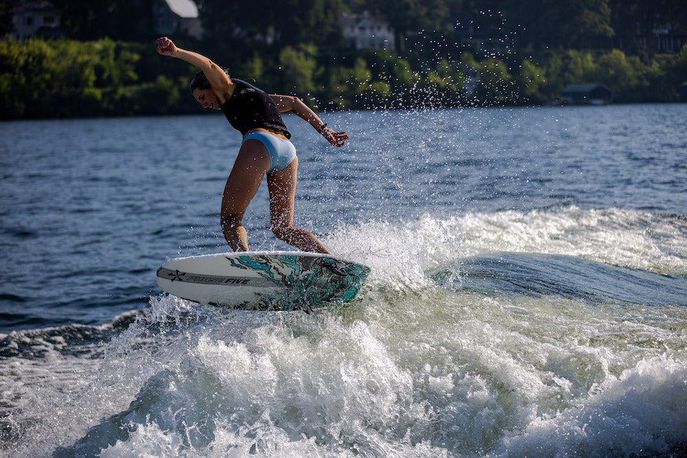 Wearing a black top and blue swim bottoms, a person rides the Phase 5 Swell Wakesurf Board 2026 by Phase 5 on a sunlit lake, with splashing water and trees in the background.