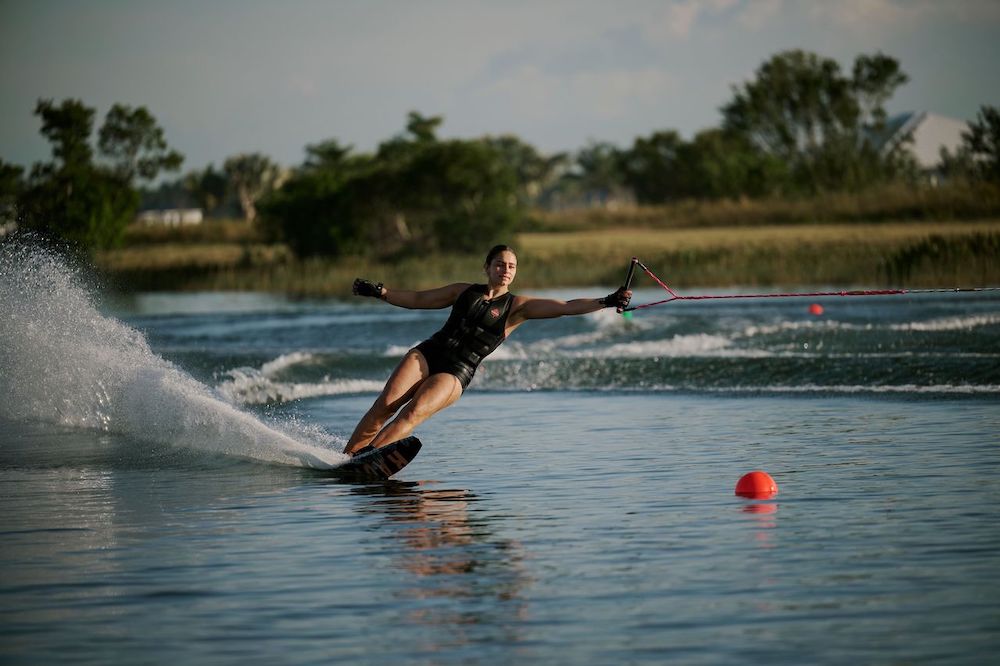 A woman in a life vest water skis on a Radar Lyric Women's Waterski | Lyric Boa + Boa ARTP 2026, leaning with a tow rope as water sprays behind her. A red buoy floats nearby, with trees and grassy banks in the background. Brand: Radar.
