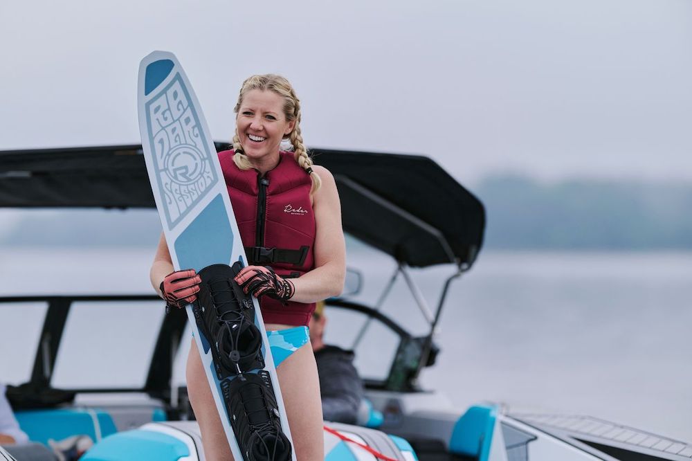 A smiling woman in a maroon life jacket holds the Radar Session Women's Waterski | Prime + ARTP 2026 by Radar while standing on a boat, with open water and an overcast sky in the background.