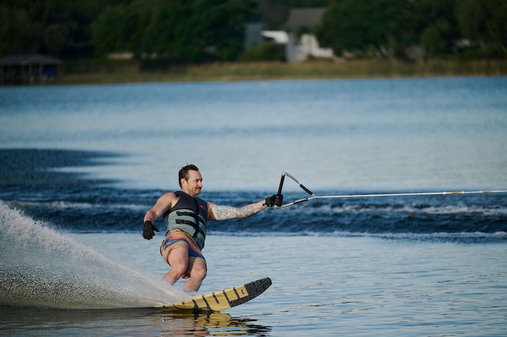 A man wearing a life vest and shorts waterskis on a lake using the Radar Union Waterski | Prime + ARTP 2026 by Radar, holding a tow rope as water sprays behind him, with trees and houses visible in the background.