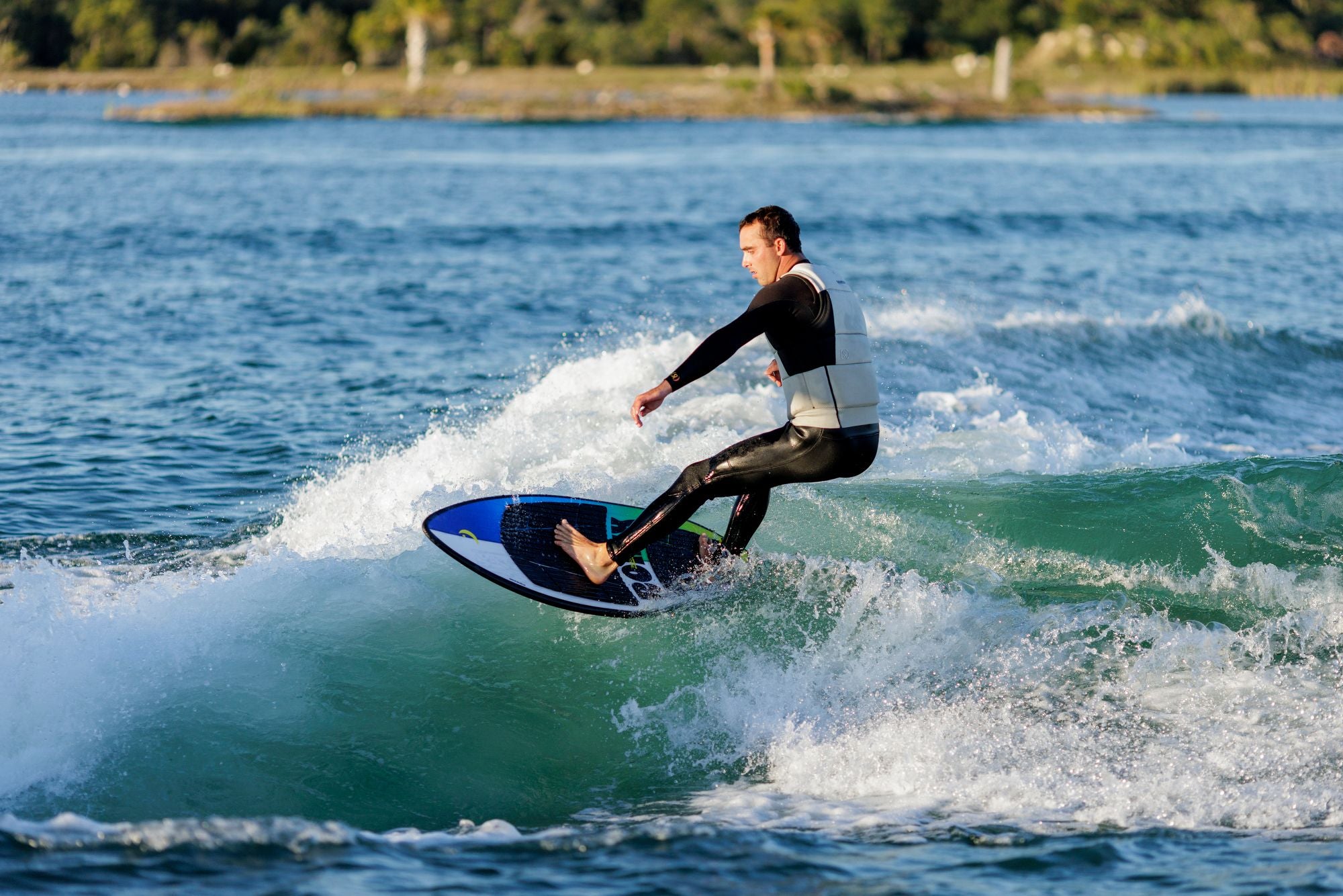 Clad in a wetsuit, an individual revels in a thrilling wakesurfing adventure, effortlessly riding the Ronix 2025 Brightside Wakesurf Board equipped with foot straps across shimmering waters surrounded by trees.