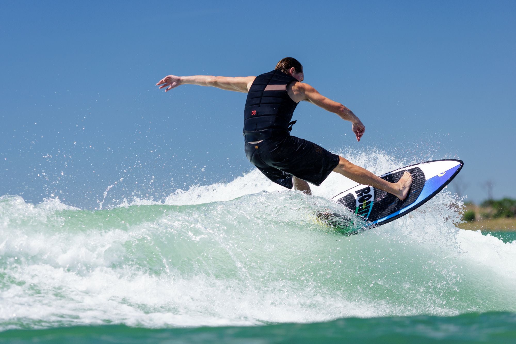 A person is wakesurfing on a wave, and their black life vest and shorts are catching the sunlight under the clear blue sky. Equipped with nimble foot straps, they glide effortlessly on their Ronix 2025 Brightside Wakesurf Board, showcasing its versatile shape on the water.