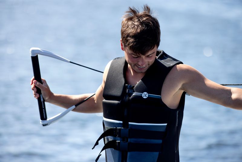 A person wearing a Ronix 2025 Covert Men's CGA Vest grips a water sport handle with splashes of water in the background.