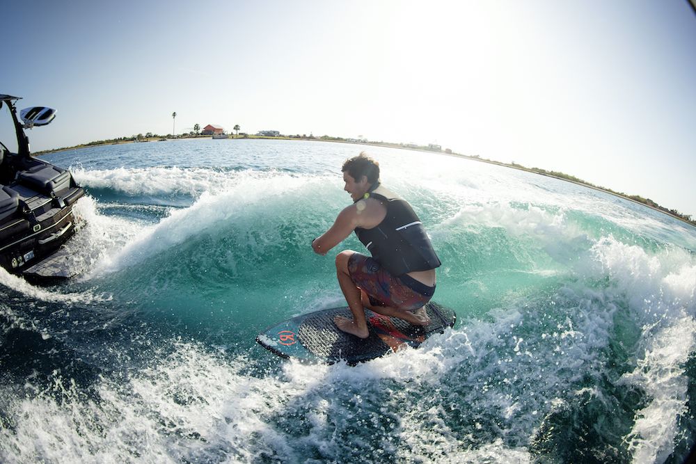 A person is wakeboarding on a wave created by a moving boat in open water under a clear sky, wearing the Ronix 2025 Covert Men's CGA Vest from Ronix, which is approved by the US Coast Guard.