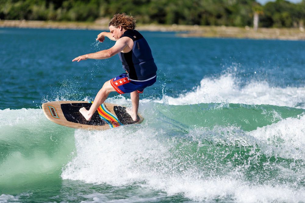 A person riding the Ronix 2025 Element Core Blunt Nose Skimmer effortlessly glides over a wave, wearing a life vest and colorful shorts, surrounded by greenery in the background.