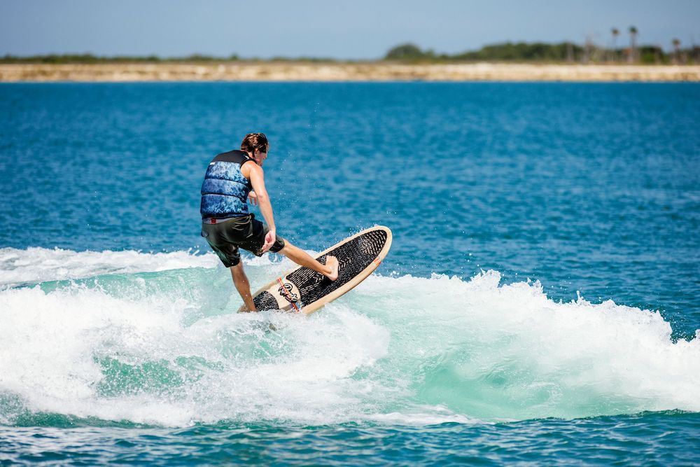 A surfer glides across the wave with effortless grace, their Ronix 2025 Element Core Longboard Wakesurf Board slicing through the ocean under a clear blue sky. The shoreline stretches in the background, echoing the harmonious surf style that seamlessly blends innovation and tradition.