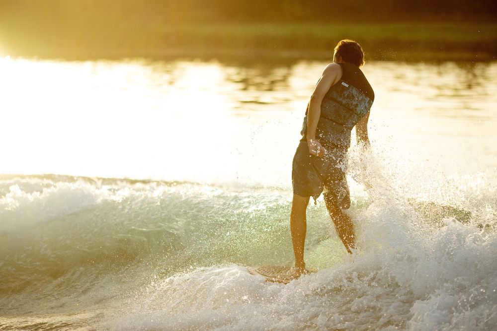 As the sun sets, a person gracefully navigates the waves exuding a relaxed surf vibe, wearing their life vest and shorts while deftly steering a Ronix 2025 Element Core Longboard Wakesurf Board, expertly made from top-quality Paulownia wood.