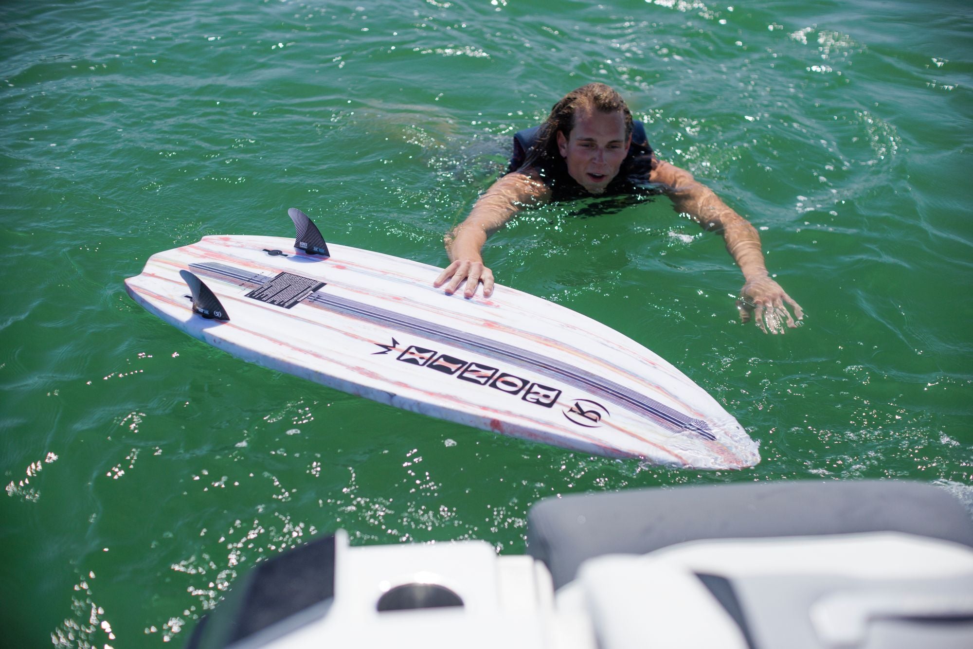 A high-performance surfer in the water reaches for the Ronix 2025 Flyweight Atlantik Wakesurf Board floating nearby in a clear, green sea.