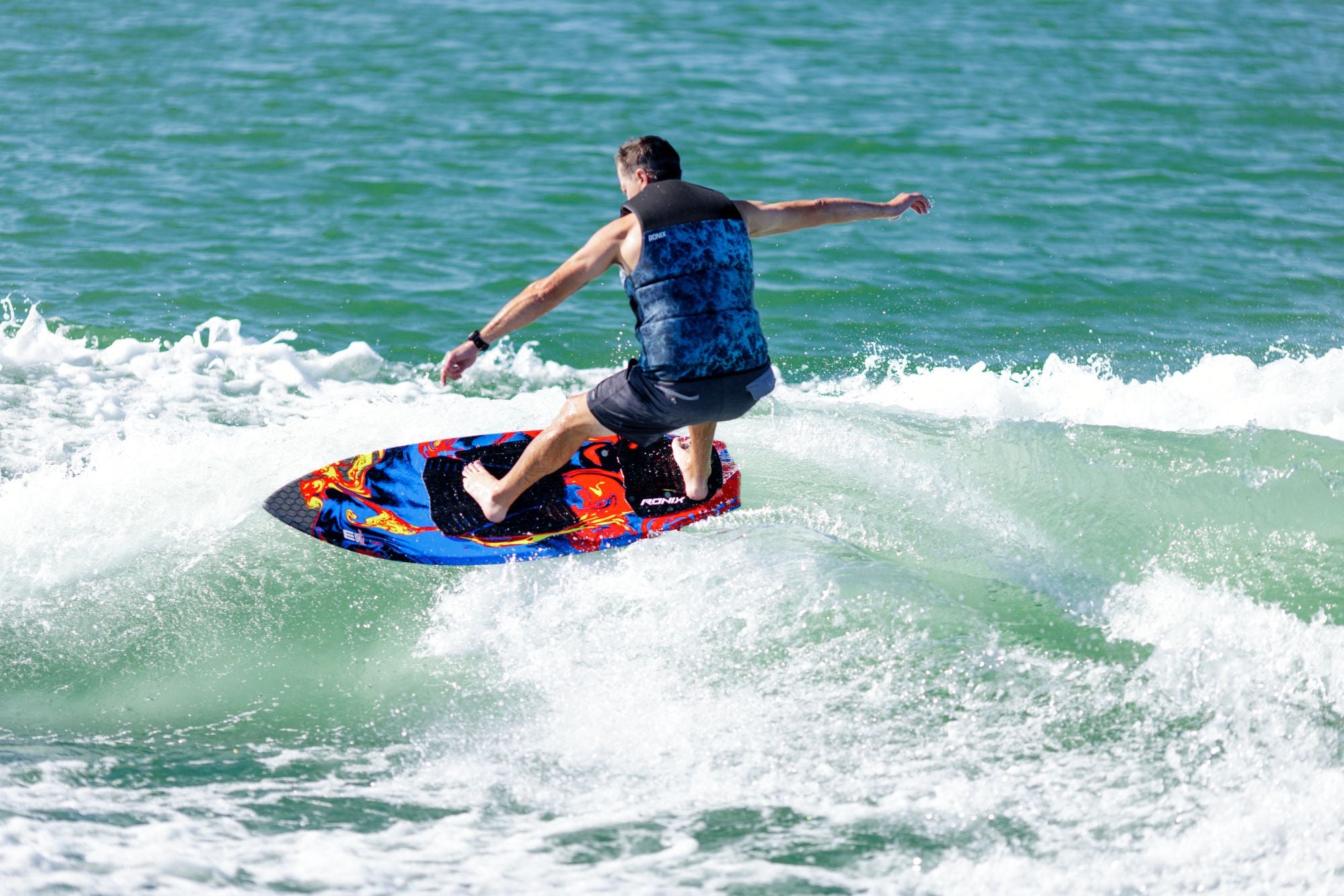 A person in a blue and black vest surfs on the Ronix 2025 H.O.M.E. Carbon Pro M50 Wakesurf Board, featuring a vibrant and versatile design, as they navigate a wave in the ocean.