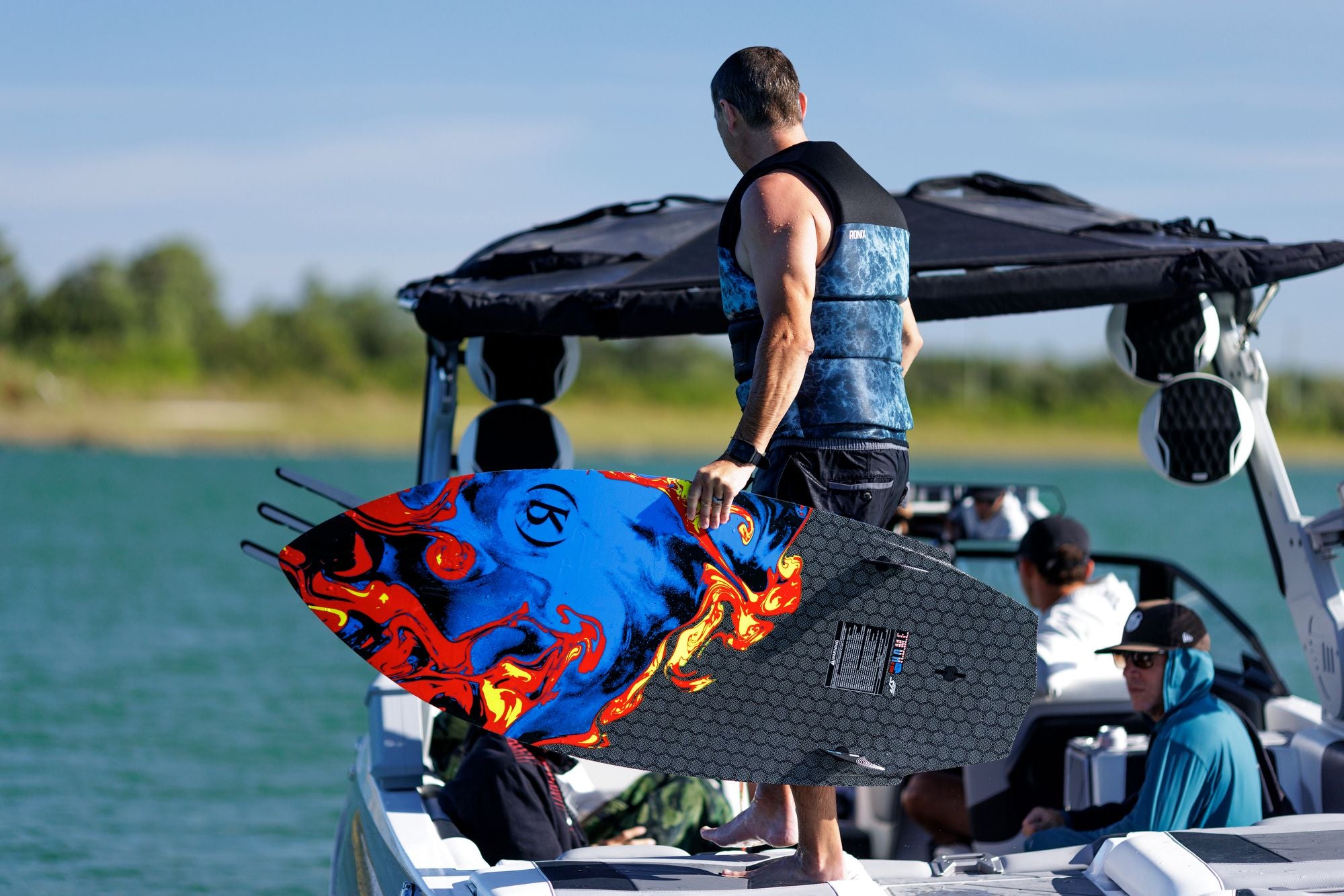 A man in a life vest grasps the Ronix 2025 H.O.M.E. Carbon Pro M50 Wakesurf Board on a boat, poised to enter the water.