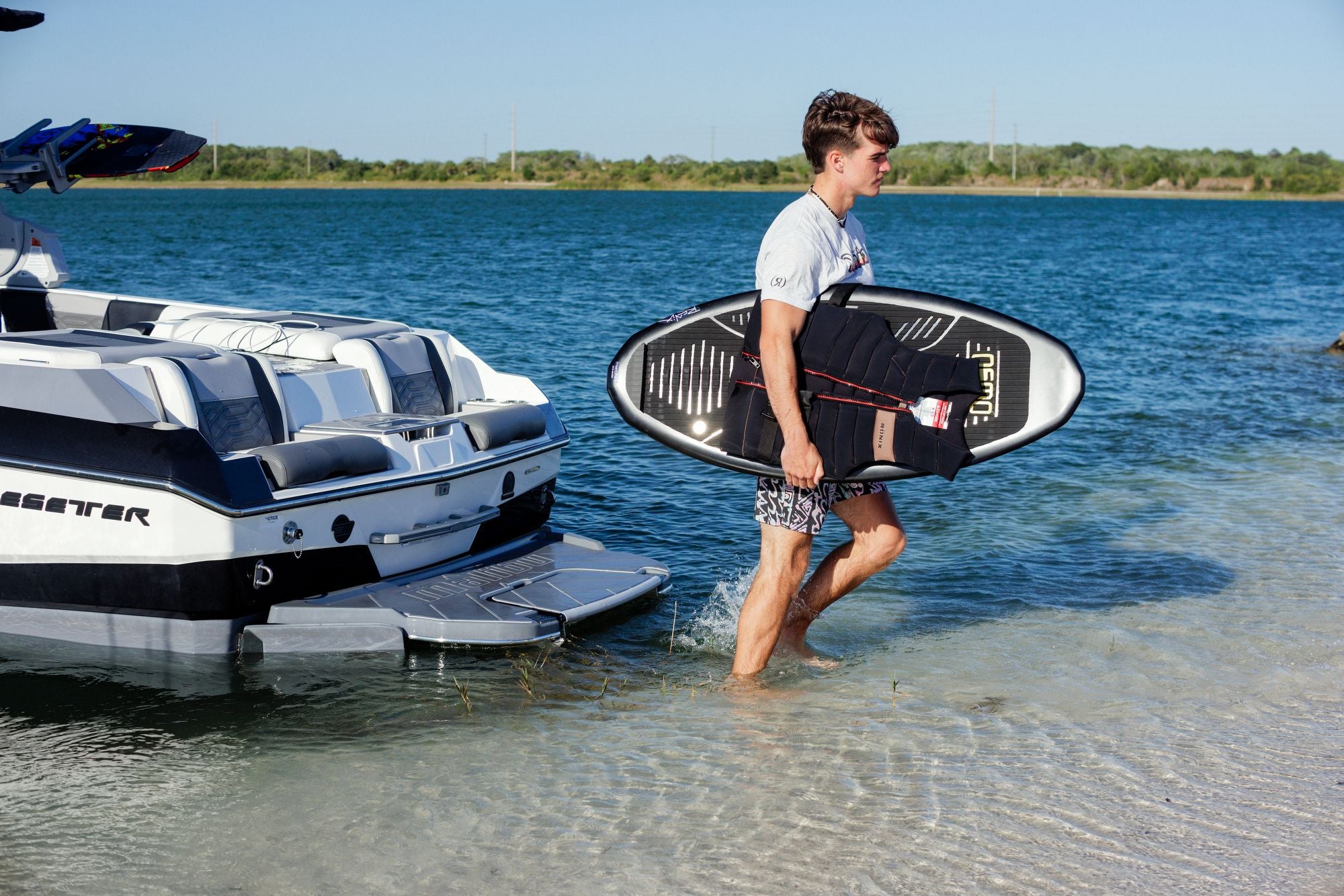 A person walks on the beach carrying a Ronix 2025 H.O.M.E. Carbon Pro Osmo Wakesurf Board, passing near a boat on the water.