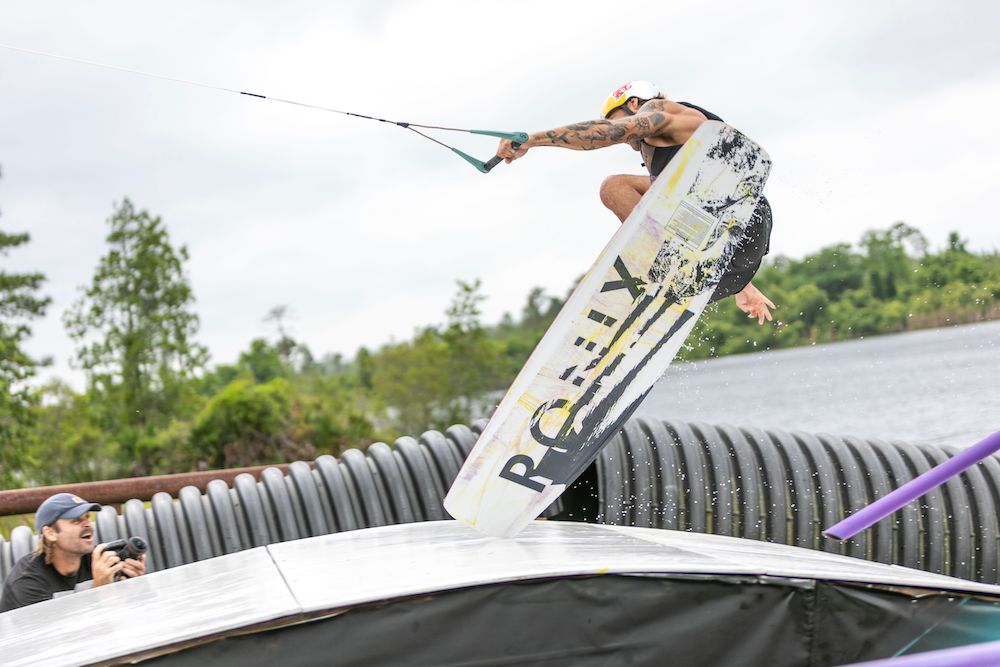 A man wearing a helmet performs a wakeboarding trick over a ramp, holding onto his Ronix 2025 Kinetic Project Wakeboard. A photographer captures the moment nearby as trees and deep channels of water form the background.