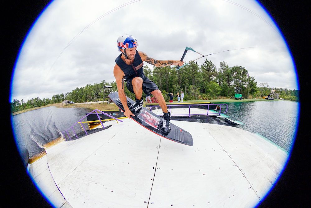 A person is using the Ronix 2025 Kinetic Project Wakeboard to perform a trick on a ramp over the lake, all beneath a cloudy sky.