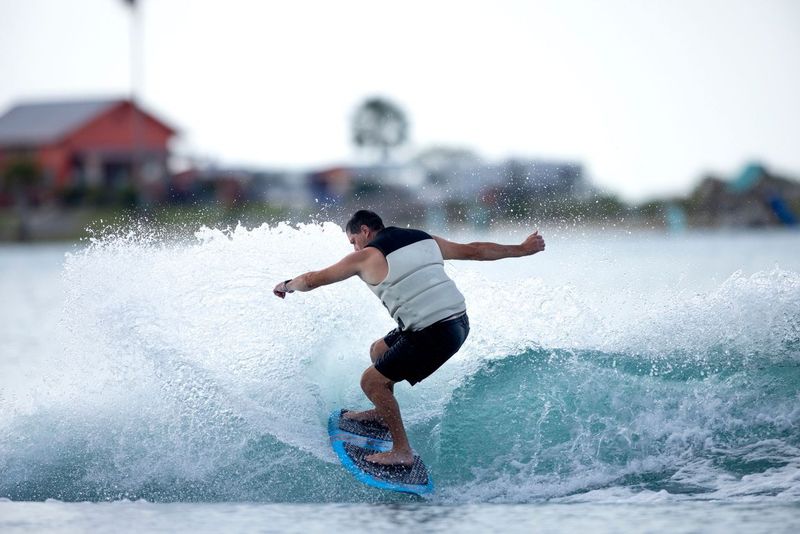 A person skillfully gliding on a wave with the finesse of wakesurfing, wearing a black and white wetsuit. The blurred buildings and trees in the background enhance the thrill, while their Ronix 2025 Marsh "Mellow" Thrasher Wakesurf Board ensures smooth maneuverability.