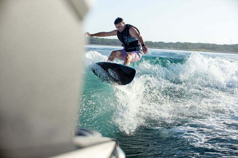 A person wakesurfing on a wave with a Ronix 2025 Marsh"Mellow" Thrasher Wakesurf Board, wearing a life vest with a lightweight soft exterior, while the distant shoreline stretches in the background.