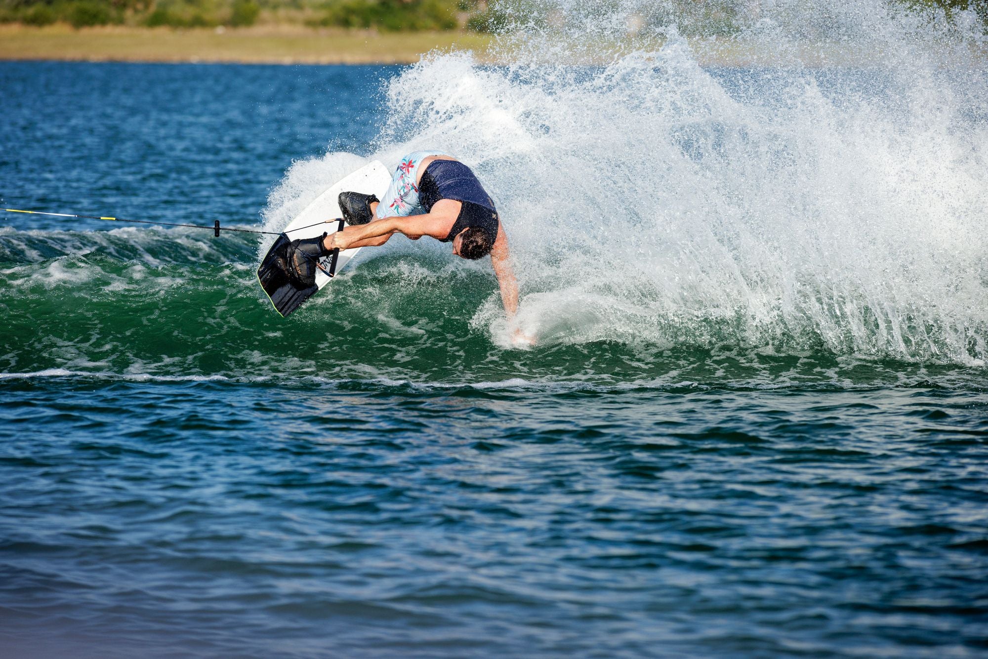 A person wakeboarding on a lake, partially submerged in water with a large splash, skillfully maneuvers with the high-response Ronix 2025 One Carbitex Boots. Gripping onto the rope, their movements are swift and precise, aided by the comfort of an Intuition+ heat moldable liner.