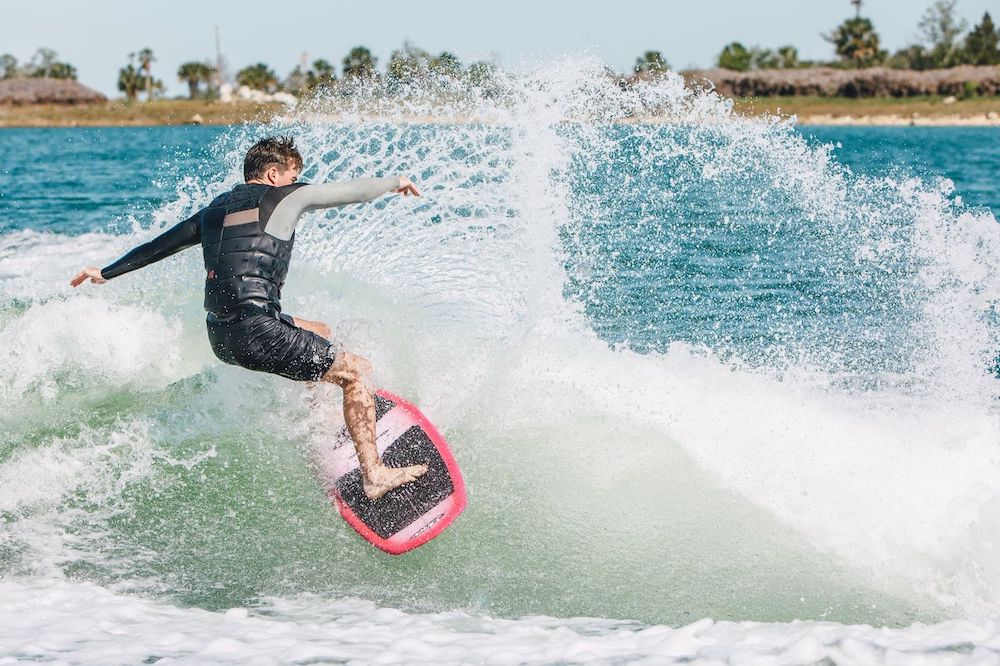 A competition-ready surfer rides a wave on the Ronix Flyweight Pro DNA Wakesurf Board 2026, sending up a spray of water. The blue ocean and green shoreline make the perfect setting to showcase Ronix’s high-performance board.