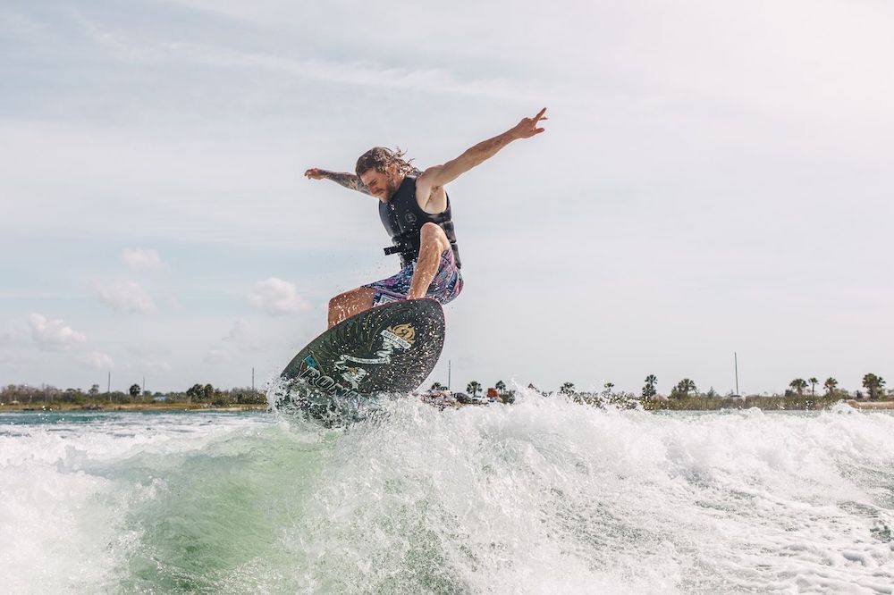 A person in a life vest and patterned shorts surfs mid-air above the water on a Ronix M50 Dark Matter Wakesurf Board 2026, arms outstretched with palm tree-lined shoreline visible in the background.