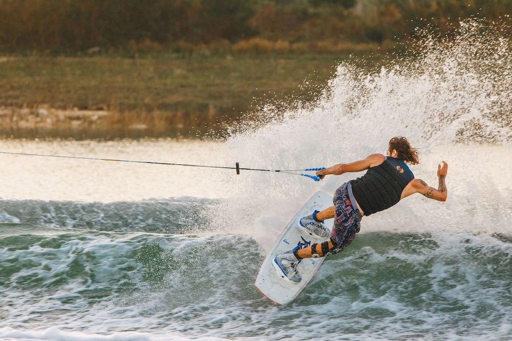 Wearing a life vest and colorful shorts, a wakeboarder in Ronix RXT Boots 2026 from Ronix leans sharply, kicking up a spray of water while gripping the tow rope, with grassy land and calm water in the background.