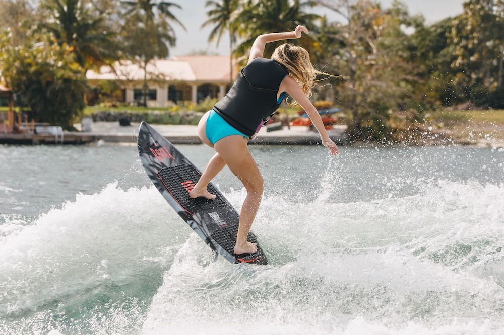 A woman in a black vest and blue shorts rides the Ronix Women's Sea Captain Wakesurf Board 2026 on a wave, water splashing around her. Trees and a house with a red roof appear in the background.