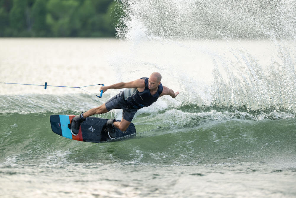 The man effortlessly glides across the lake, expertly maneuvering his Hyperlite 2024 Murray Pro Wakeboard with the precision of a seasoned rider. With a Biolite 3 Core providing unparalleled strength and responsiveness, and