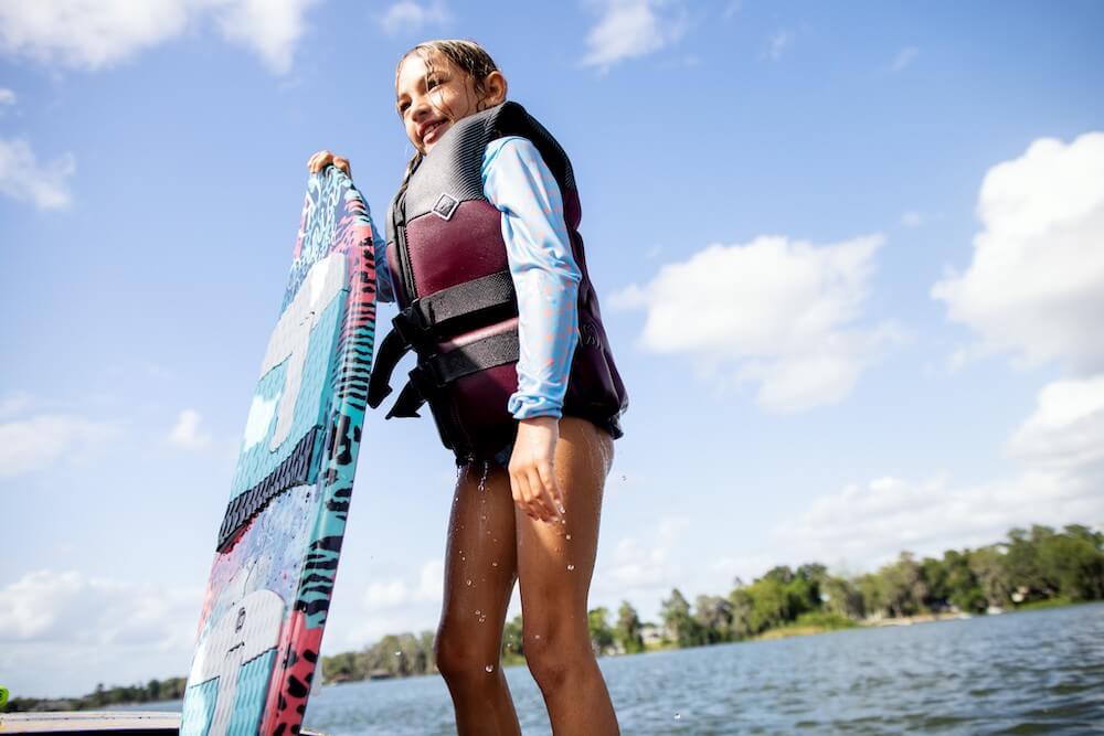 A young girl standing on a boat holding a wakeboard while wearing the Ronix Atlantis Capella 3.0 Junior CGA Vest.