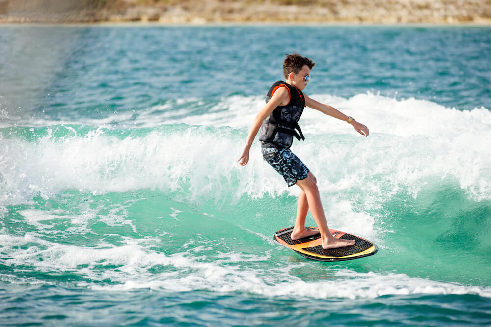 A young boy riding a wave on a surfboard, showcasing the Ronix Neptune Capella 3.0 Junior CGA Vest sizing size.