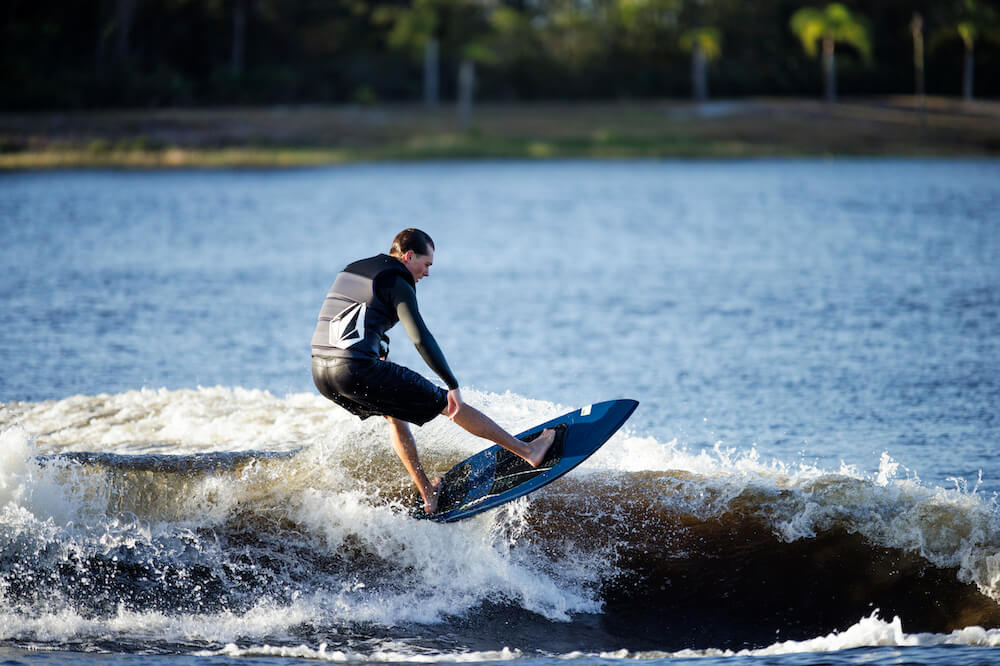 A man riding a wave on a Ronix 2024 H.O.M.E. Carbon Pro M50 Wakesurf Board equipped with a wakesurfer and button fin system provided by Ronix.
