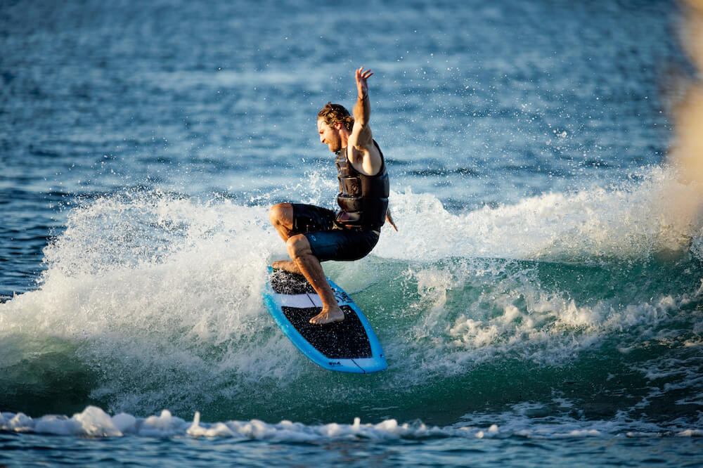 A man is riding a wave on a Ronix 2024 Flyweight Pro DNA Wakesurf Board.