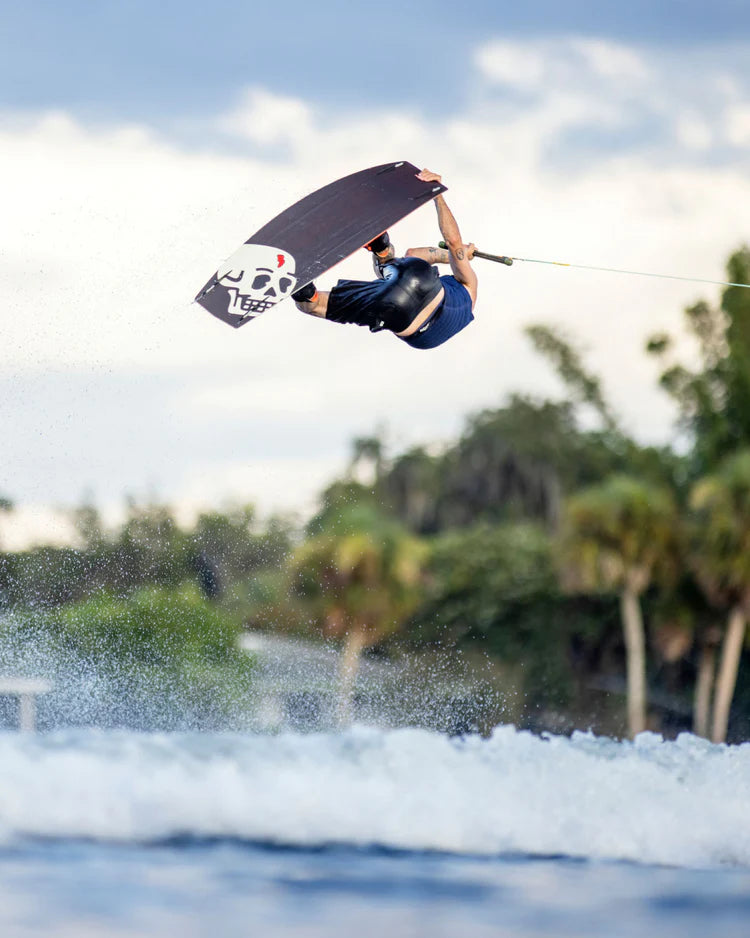 A man gracefully performing wakeboarding stunts in the air over a serene lake, experiencing the comfortable sensation of impact protection and relying on Follow Wake's Follow 2022 BP Pro Men's Jacket - Navy.