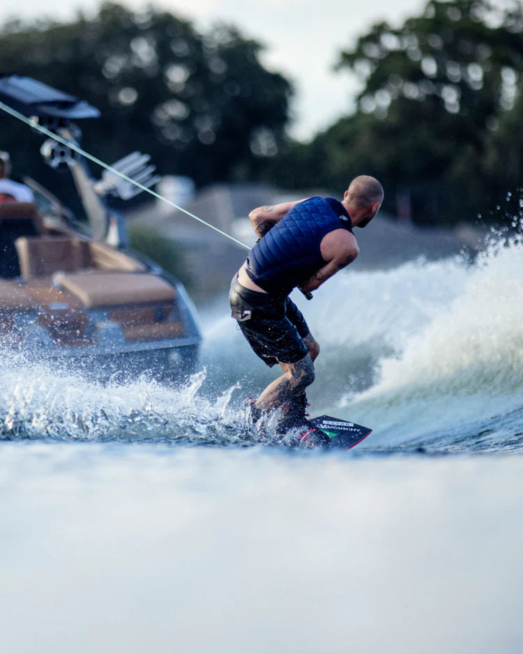 A man wakeboarding on a lake with a boat behind him, experiencing comfortable and durable impact protection wearing the Follow 2022 BP Pro Men's Jacket - Navy by Follow Wake.