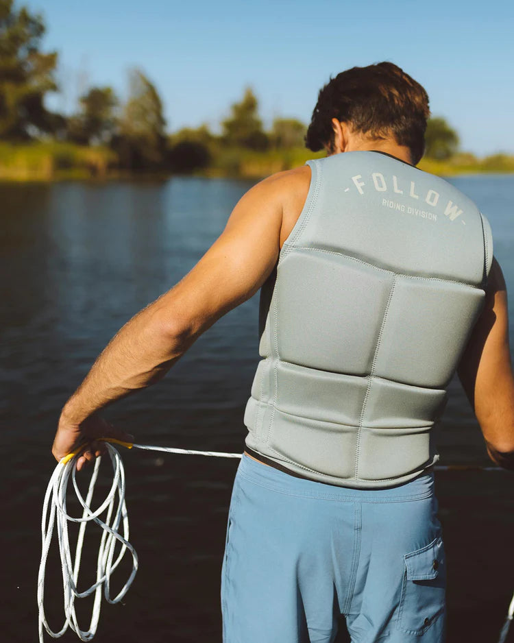 A man wearing a Follow Wake 2022 Division Men's Jacket - Stone with dual layer construction and segmented panels on a body of water.