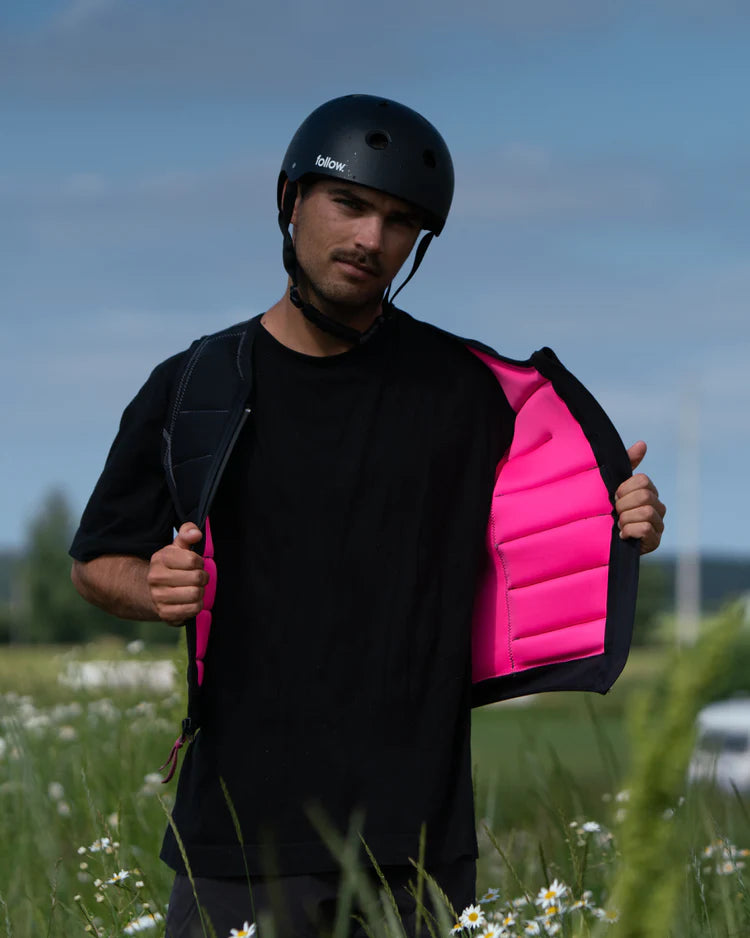 A man wearing a Follow 2022 TBA Men's Jacket - Black by Follow Wake and a pink vest standing in a field.
