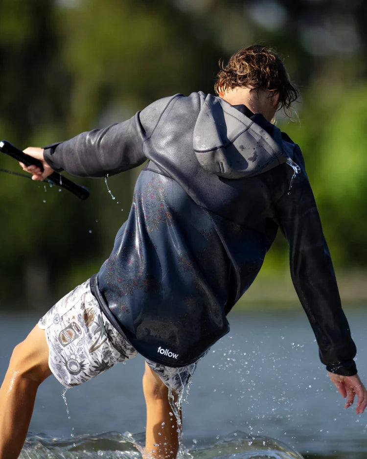 A man is riding a wakeboard in the water, wearing a Follow 3.12 Anorak Pro Neo Outer Jacket - Charcoal by Follow Wake to combat the wind chill factor.