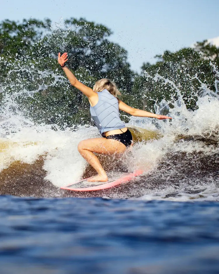 A woman riding a wave on a surfboard, wearing a Follow Atlantis Ladies Jacket - Ash Blue by Follow Wake.