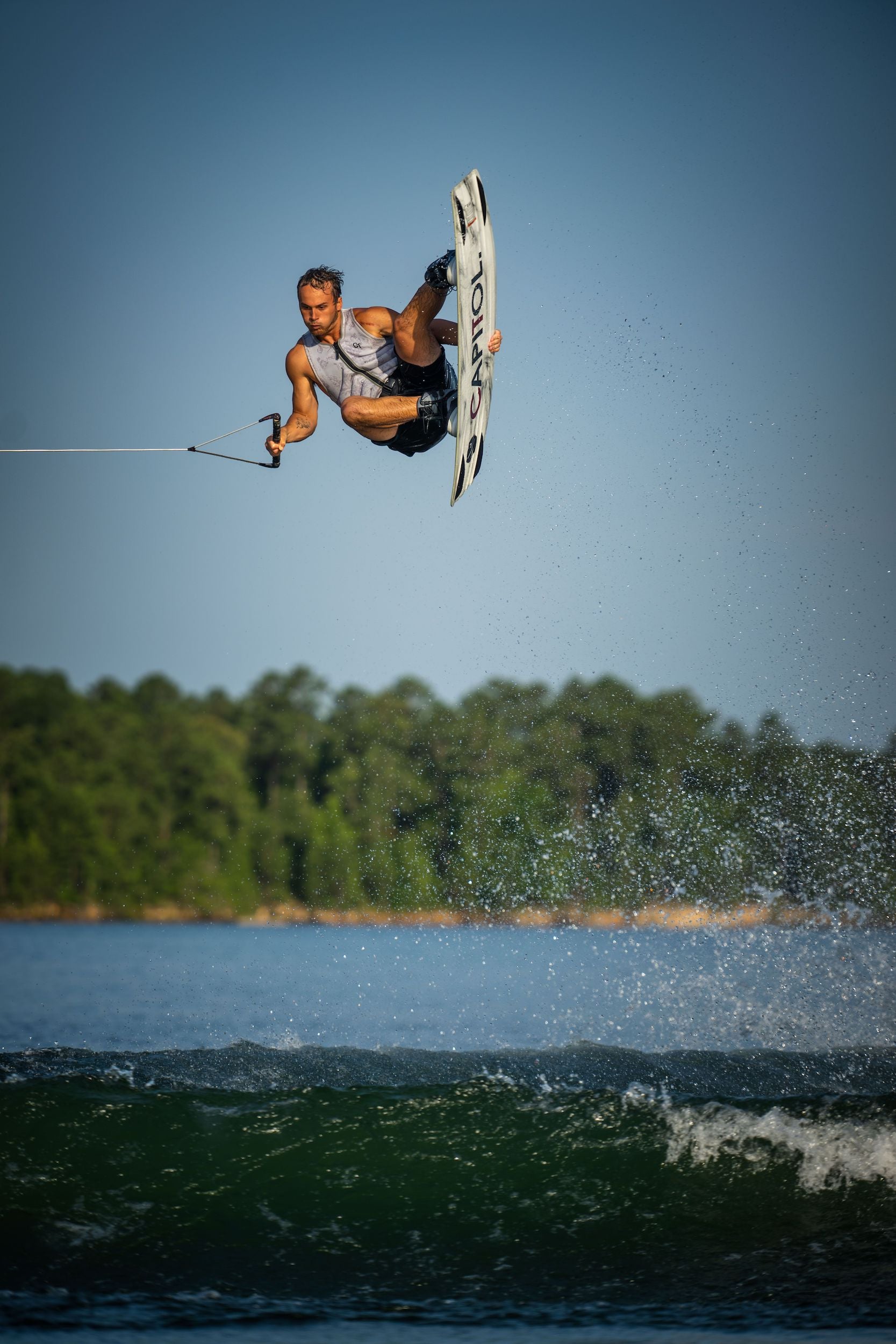 Cory Teunissen, a professional wakeboarder, gracefully soars through the air while wakeboarding over a picturesque lake on the Hyperlite 2023 Capitol Wakeboard by Hyperlite.
