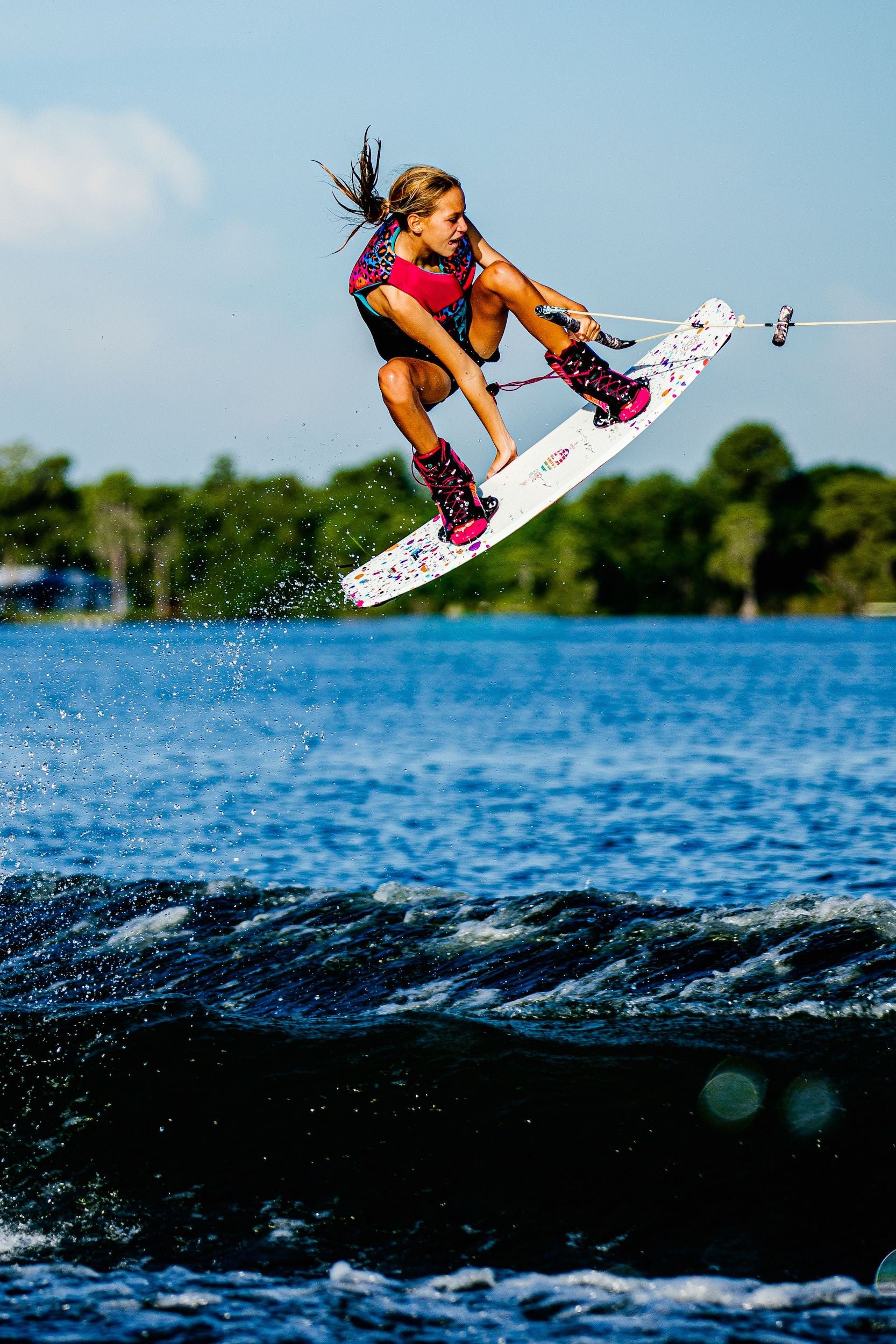A woman is gracefully wakeboarding in the air, showcasing her skills on a Hyperlite Shaun Murray Signature Grom Model. She exudes confidence and enthusiasm, embodying the spirit of Lil It Girls in Hyperlite 2024 Murray Girls Wakeboard.