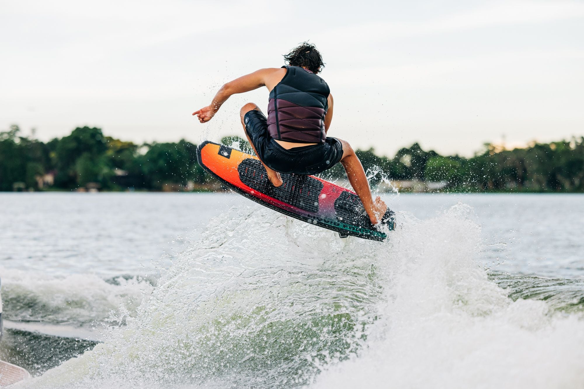 A man performing high-performance wakeboarding tricks on a lake using the Liquid Force 2024 Pod Wakesurf Board.