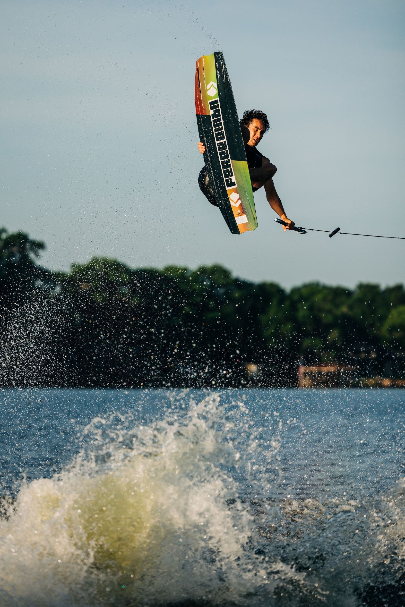 A man wakeboarding in the air over a body of water while performing tricks and riding a Liquid Force 2023 Rhyme Wakeboard with a 3-Stage Rocker, displaying the skill and determination of professional wakeboard