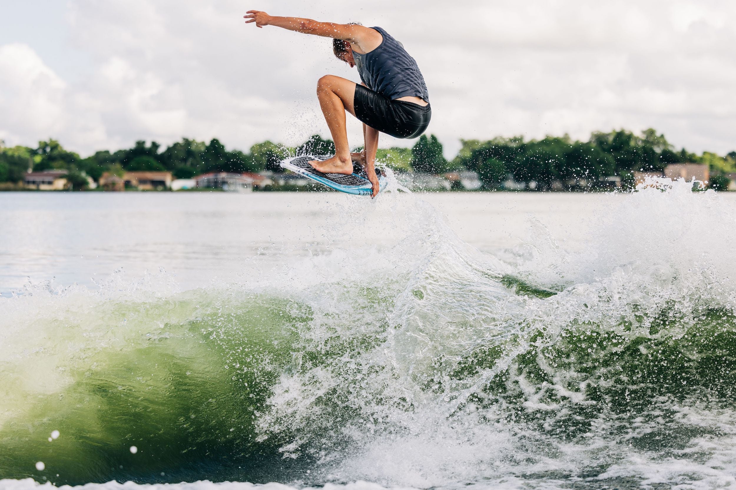 A man is effortlessly riding a powerful wave on his Liquid Force 2024 Twinzer Wakesurf Board, expertly maneuvering with the assistance of a Twinzer fin configuration. As he glides along the water's surface, the