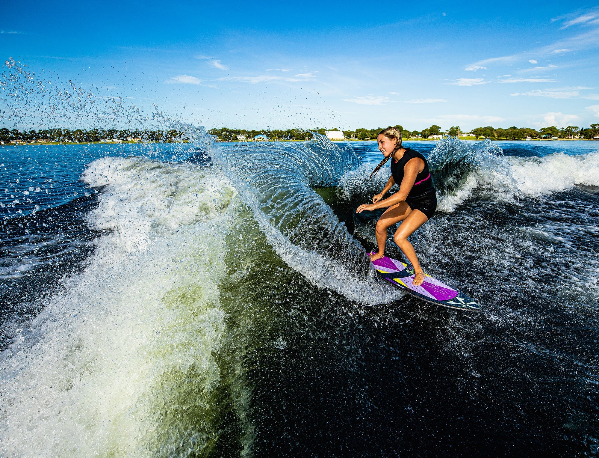 A woman riding a wave on a surfboard, showcasing her impressive skills in skim style. She confidently maneuvers on the Phase 5 2023 Diamond Luv Wakesurf Board, which features advanced construction materials such as Carbon Inn.