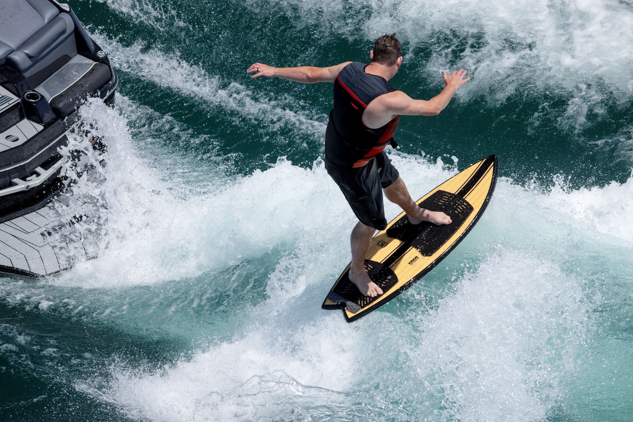 A man showcases his versatility as he rides a Ronix 2024 Koal Classic Fish Wakesurf Board on top of a boat, combining the thrill of wakesurfing with the punchy waves.