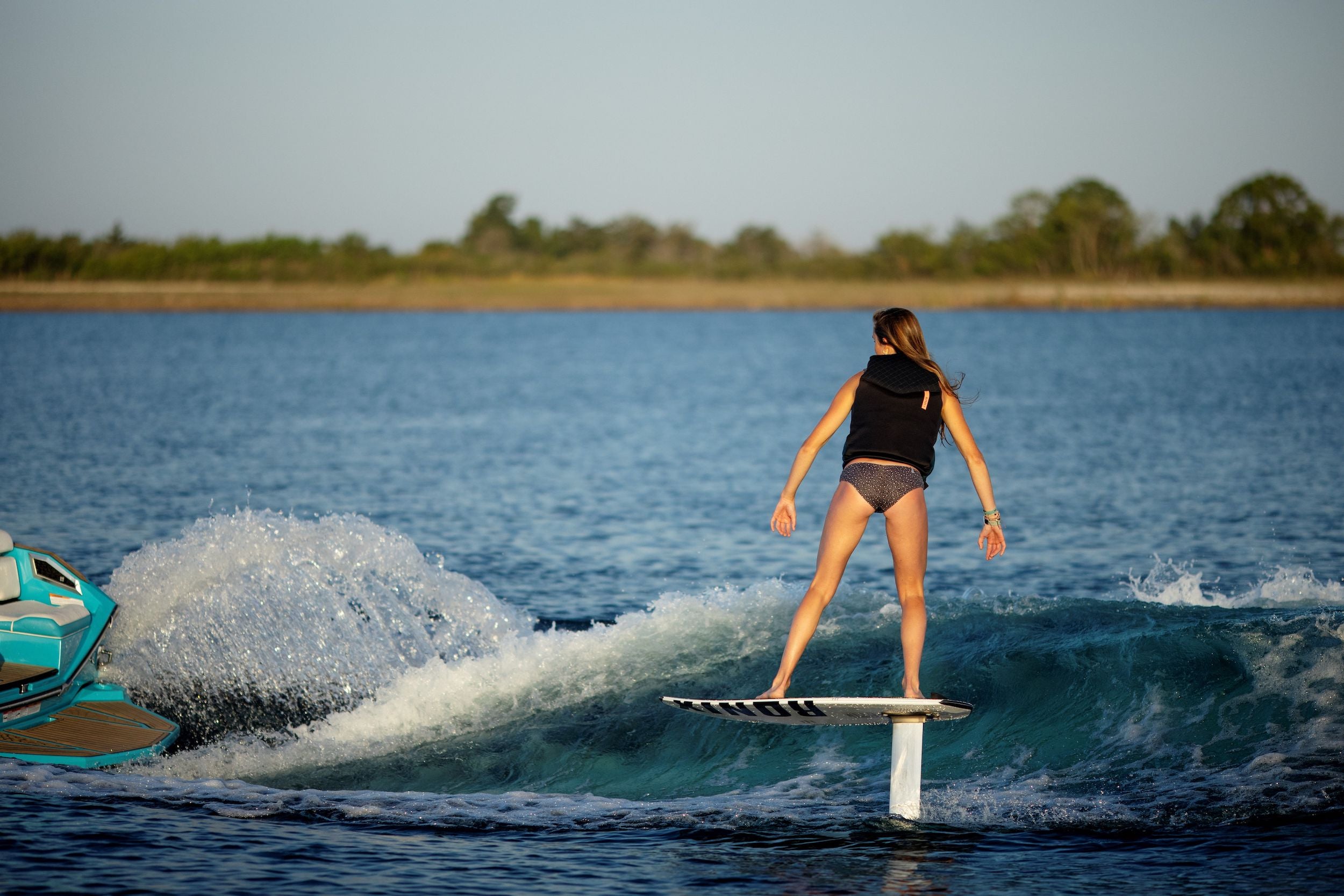 A woman is standing on a surfboard in the water, confidently showcasing the water-resistant features of the Ronix Women's Imperial Capella 3.0 CGA Vest model.